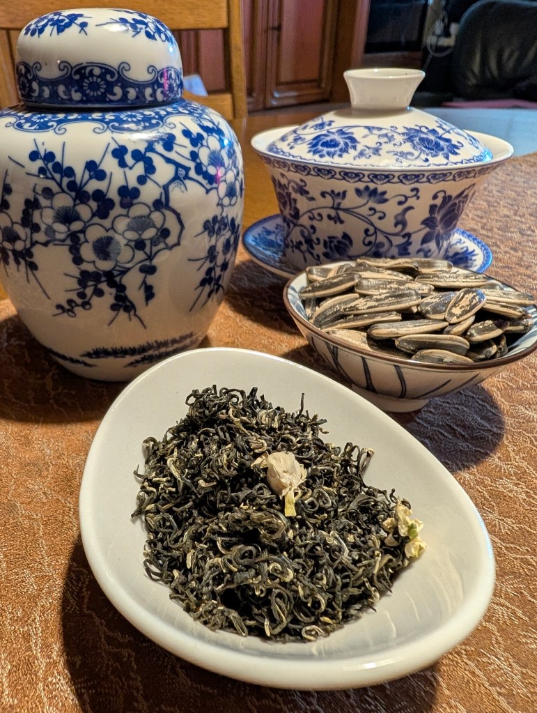 A close-up view of a decorative tea jar, a tea bowl, and loose tea leaves presented in a white dish, accompanied by sunflower seeds, set on a textured surface.