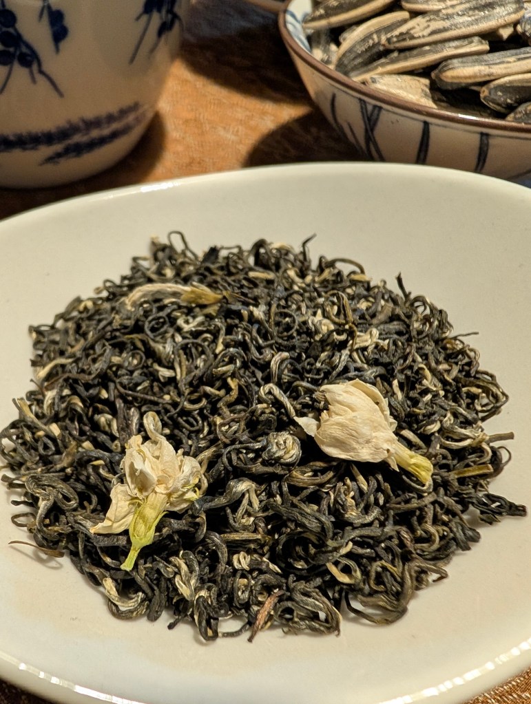 A close-up of loose Jasmine tea leaves with dried jasmine flowers on a plate, accompanied by a traditional tea cup and a bowl of sunflower seeds in the background.