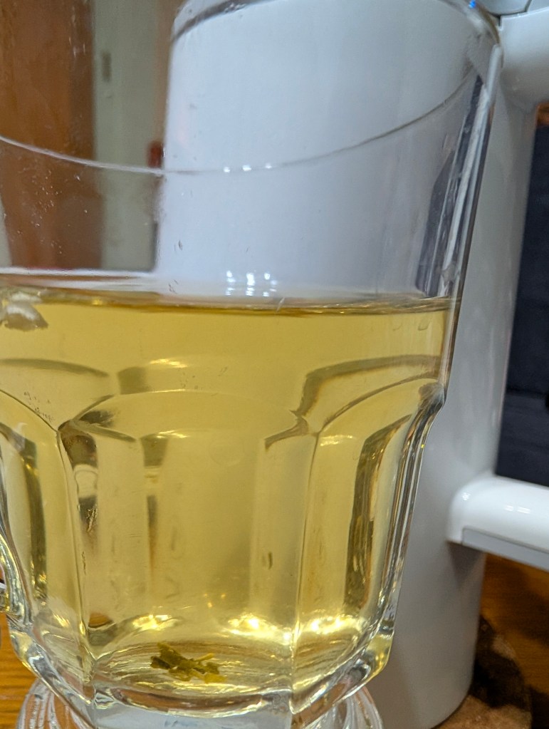A clear glass filled with lightly colored brewed tea, showcasing a delicate floral aroma. The glass is positioned on a wooden surface with a white teapot in the background.