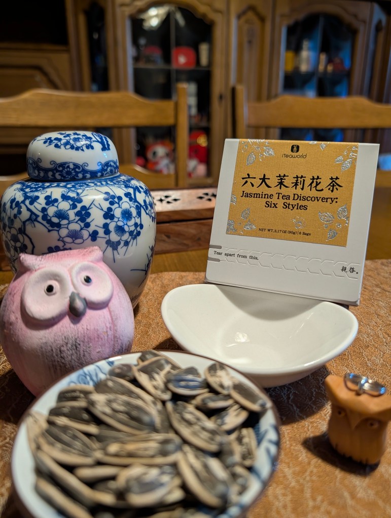 A decorative blue and white ceramic tea jar next to a pink owl figurine and a white dish, accompanied by a bowl of sunflower seeds and a card detailing the Jasmine Tea Discovery box from iTeaworld.