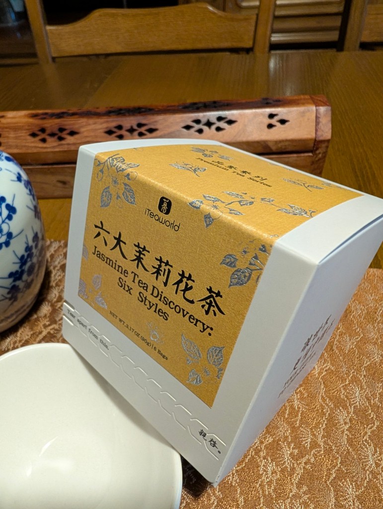 A Jasmine Tea Discovery box from iTeaworld, featuring a gold and white design, placed beside a tea bowl on an intricately patterned tablecloth.
