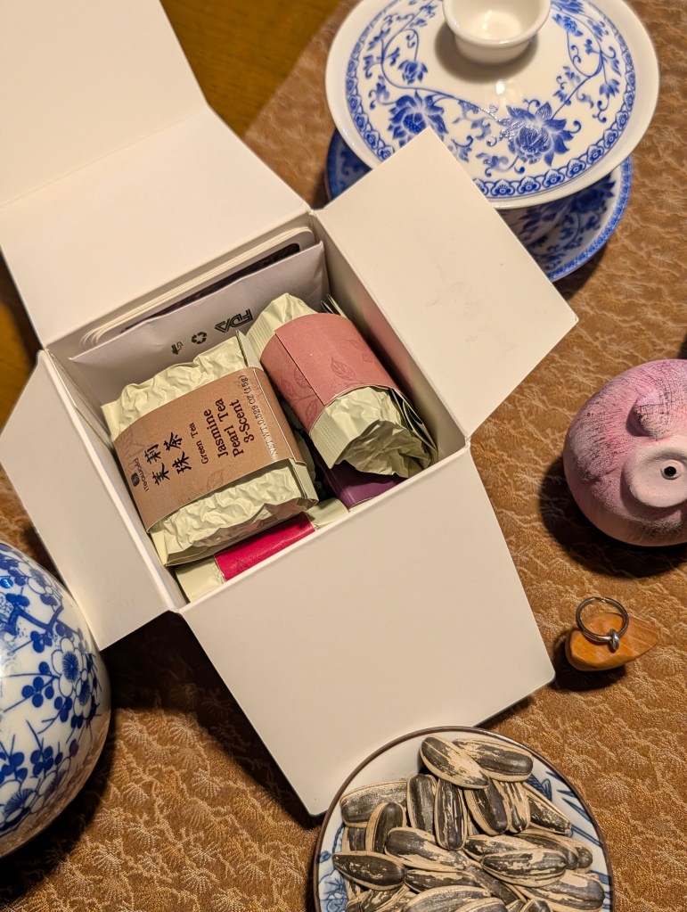 An open white box displaying several packages of Jasmine tea, surrounded by traditional tea ware and a plate of sunflower seeds.